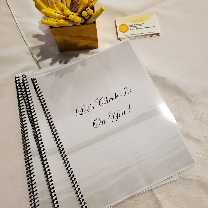 photo of three booklets stacked on a white tablecloth. A small gold box with yellow tipped pens. A stack of business cards with yellow logo and design are in the upper right corner.